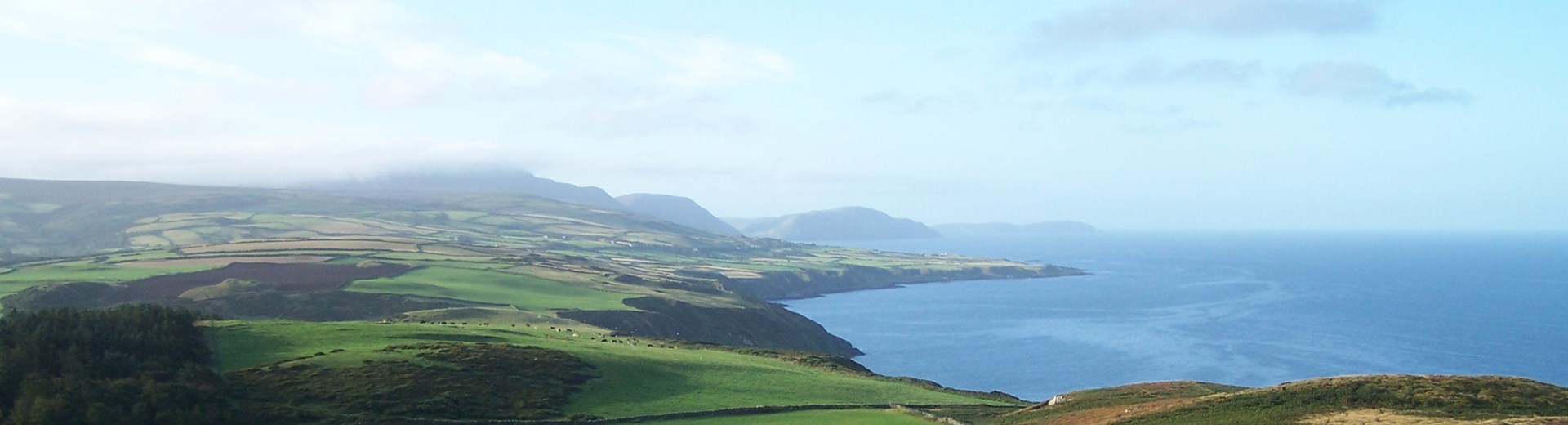 The Isle Of Man taken from above. Green rolling hills and a coastline with blue skies. 